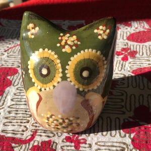 May include: A ceramic owl figurine with a green body and large yellow and white eyes. The owl has a light purple nose and is decorated with small, colorful dots. The owl is a decorative item.