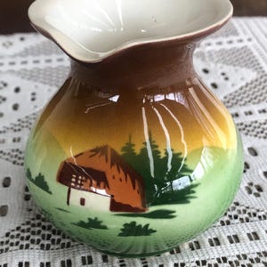 May include: A small, ceramic pitcher with a landscape design. The pitcher features a white interior, a brown rim, and a rounded body with a scene of a house, trees, and a mountain range in shades of green, yellow, and brown.