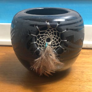 May include: A dark blue, round ceramic vase with a dreamcatcher design. The vase has etched lines and a woven dreamcatcher with a feather attached. The vase is sitting on a wooden surface.