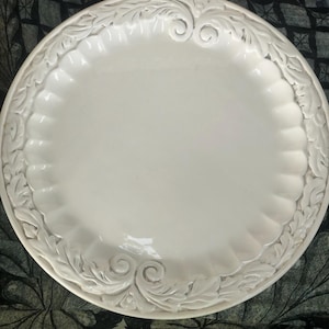 May include: A round, off-white ceramic plate featuring a smooth, flat center and a wide, scalloped rim. The border is decorated with a raised, embossed relief pattern of swirling acanthus leaves and scrolls that encircle the entire edge.