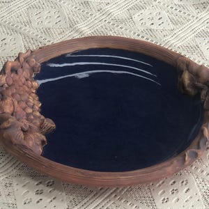 May include: A handmade ceramic serving dish with a dark blue glaze and brown sculpted handles. The handles are shaped like vines with leaves and grapes.