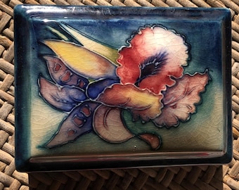 Puede incluir: Caja rectangular de cerámica con diseño floral. La caja tiene un borde azul oscuro y un fondo azul. El diseño floral presenta pétalos rojos, amarillos y morados. La caja está colocada sobre una superficie tejida.