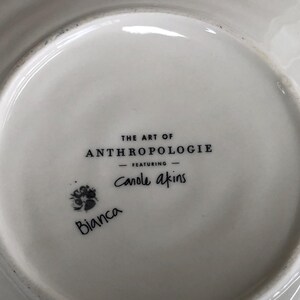May include: White ceramic plate with the text "THE ART OF ANTHROPOLOGIE" and "- FEATURING - Carole akins". A small floral design and the name "Bianca" are also visible.