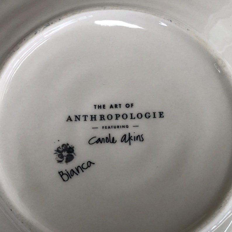May include: White ceramic plate with the text "THE ART OF ANTHROPOLOGIE" and "- FEATURING - Carole akins". A small floral design and the name "Bianca" are also visible.