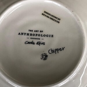 May include: White ceramic plate with the text "THE ART OF ANTHROPOLOGIE" and "Canole akins". The plate also has the text "Chipper" and a sticker that says "DISHWASHER & MICROWAVE SAFE MADE IN CHINA/FABRIQU&Eacute; EN CHINE".