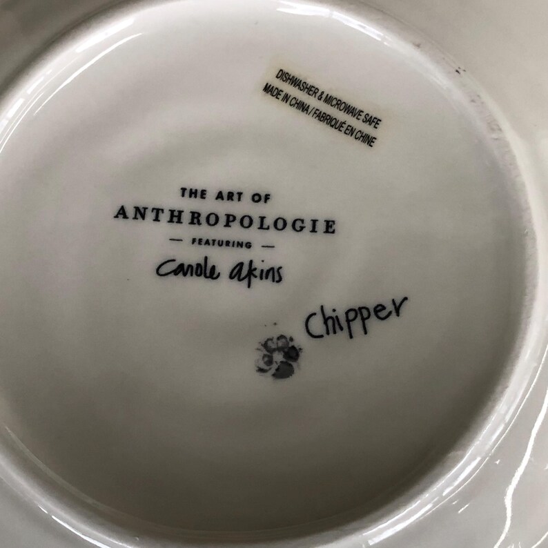 May include: White ceramic plate with the text "THE ART OF ANTHROPOLOGIE" and "Canole akins". The plate also has the text "Chipper" and a sticker that says "DISHWASHER & MICROWAVE SAFE MADE IN CHINA/FABRIQU&Eacute; EN CHINE".