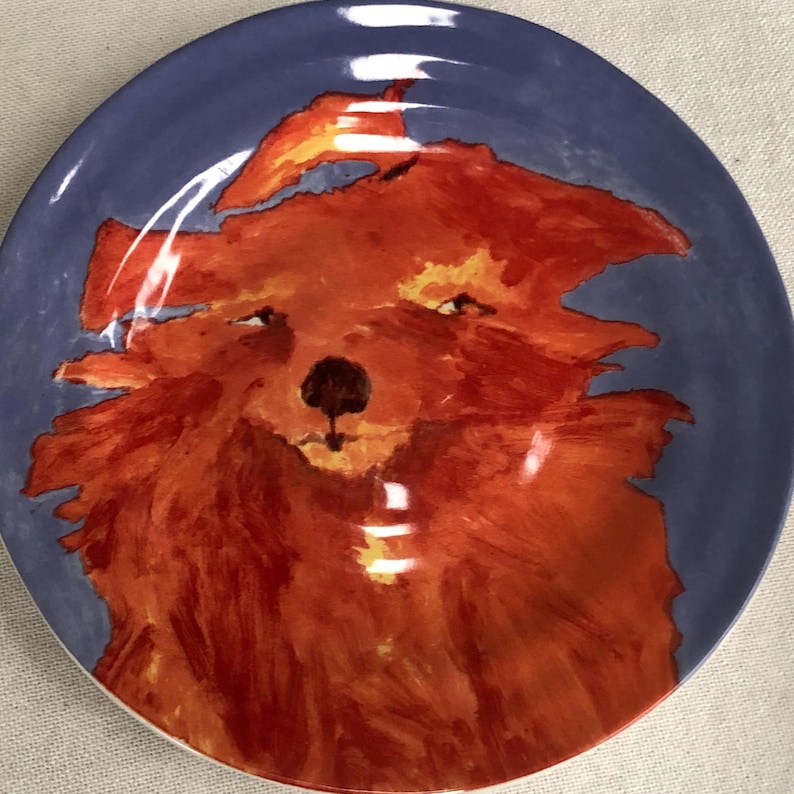 May include: A round ceramic plate featuring a hand-painted red fox design on a blue background. The fox is depicted with a fluffy texture, and the plate has a glossy finish. The plate is suitable for serving food.