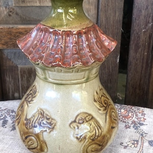 May include: A ceramic vase with a light beige body featuring embossed brown lion figures around the base. The neck is glazed in olive green, and the shoulder is decorated with a textured, reddish-brown, roof-like architectural element.