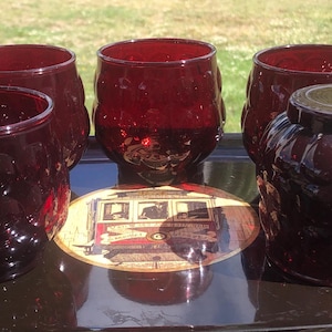 May include: A set of four deep red glass tumblers with a textured hobnail pattern. Three tumblers are upright, and one is inverted. They are arranged on a dark tray featuring a circular graphic of a vintage cable car with the text San Francisco.