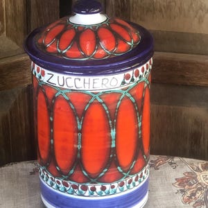 May include: A cylindrical ceramic canister with a lid, featuring a vibrant orange and blue geometric pattern. The word ZUCCHERO is printed in black lettering near the top rim. The design includes repeating oval shapes and decorative borders.