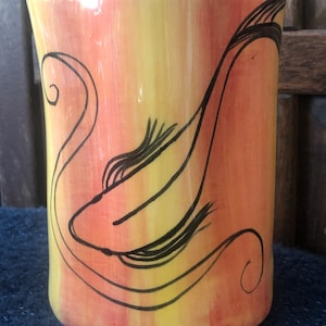 May include: A cylindrical ceramic mug with a gradient of yellow, orange, and red hues. A black line drawing of a stylized fish decorates the mug. The mug has a black rim and a white interior. The mug is on a dark blue surface.