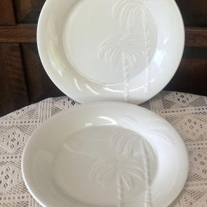 May include: Two white ceramic plates with embossed palm tree designs. The plates are round with a slightly raised edge. The palm tree design features two trees with detailed fronds and trunks. The plates are set on a white lace tablecloth.