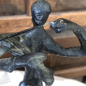 May include: A dark bronze sculpture of a person playing a stringed instrument. The figure is depicted in a seated position, with arms raised and a bow in hand. The sculpture has a textured surface and a weathered appearance.
