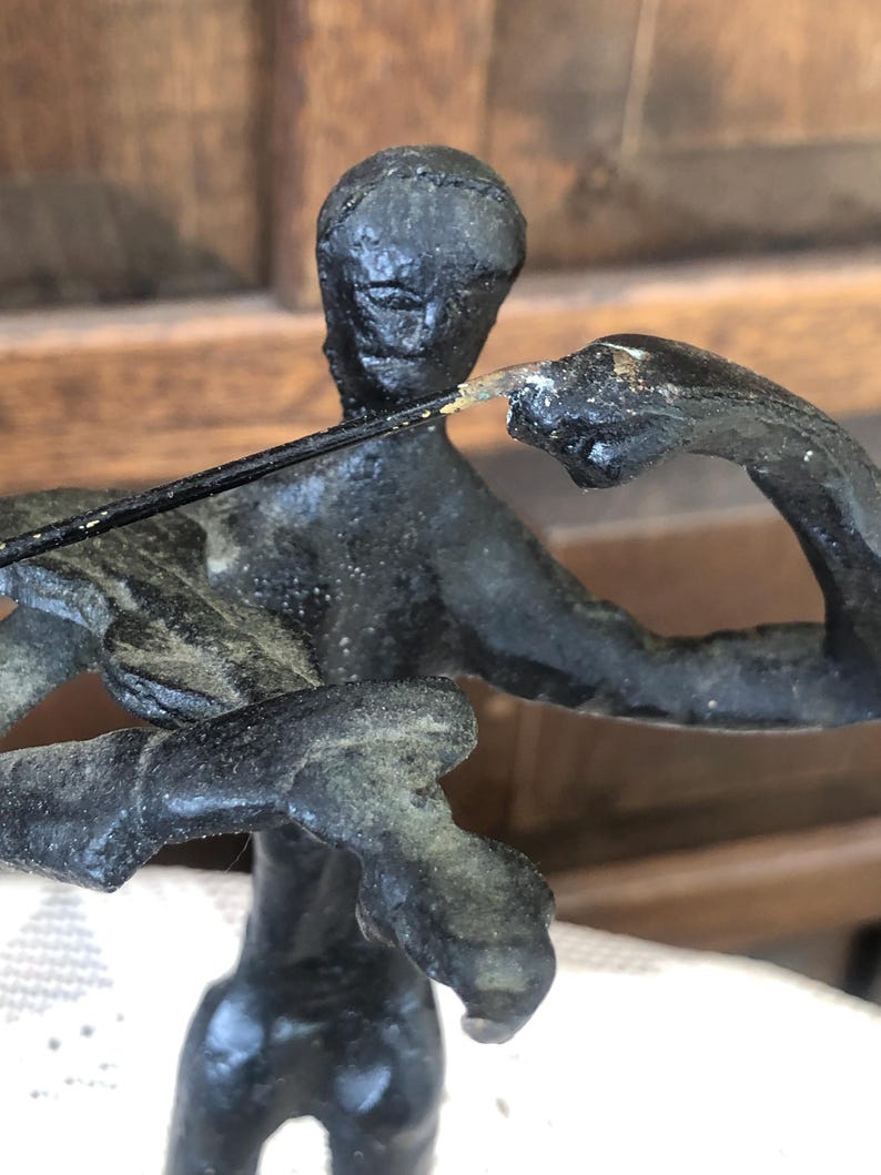 May include: A dark bronze sculpture of a person playing a stringed instrument. The figure is depicted in a seated position, with arms raised and a bow in hand. The sculpture has a textured surface and a weathered appearance.