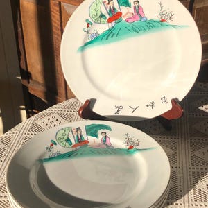 May include: White ceramic plates with a hand-painted Asian-inspired design. The plates feature a scene with figures, trees, and text in black. The plates are stacked and displayed on a table.