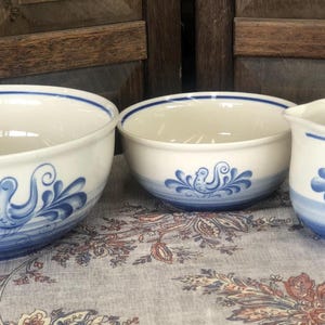 May include: A set of three ceramic kitchen items including a large mixing bowl, a smaller bowl, and a creamer pitcher. Each piece is off-white with a blue bird motif and blue horizontal bands around the base and rim of the vessels.
