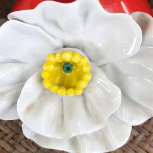 May include: Close-up of a white ceramic flower with a yellow center and a green accent. The flower has multiple layers of petals and is set against a red background. The flower is likely a decorative item.