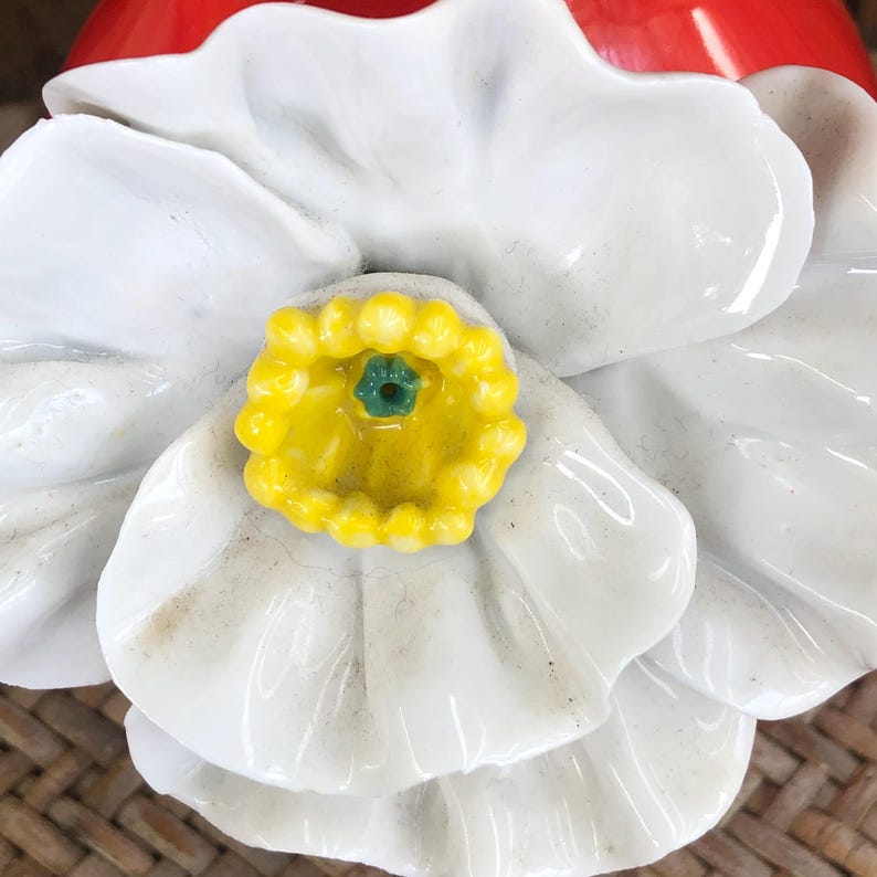 May include: Close-up of a white ceramic flower with a yellow center and a green accent. The flower has multiple layers of petals and is set against a red background. The flower is likely a decorative item.