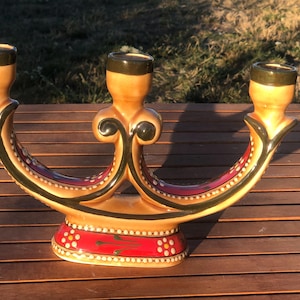 May include: A ceramic candelabra featuring three candle holders with a curved, arched base. The piece is finished in a glossy tan glaze with dark green accents, red panels, and white dot detailing along the edges and base, including small floral motifs.