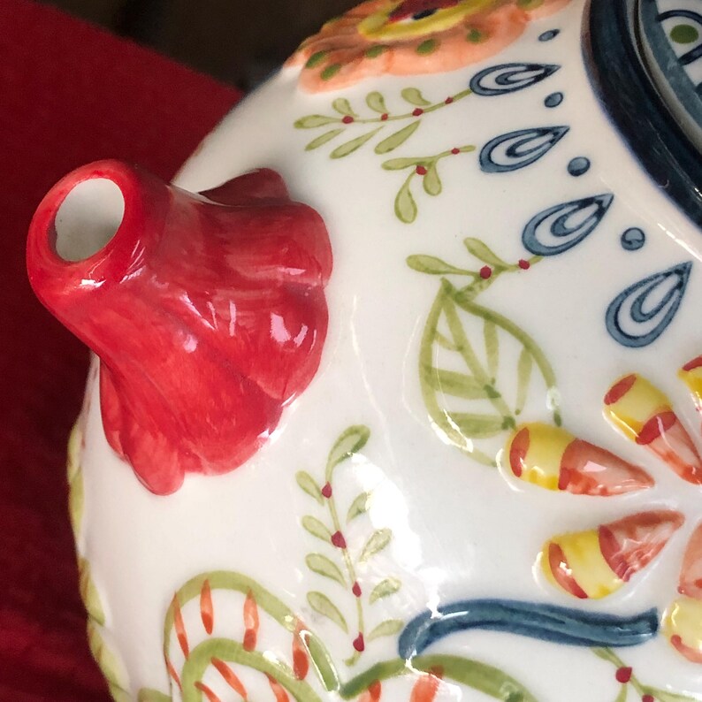 May include: Close-up of a white ceramic teapot with a red spout and handle. The teapot is decorated with colorful floral and leaf patterns in orange, blue, green, and yellow. The design is hand-painted and has a raised relief texture.
