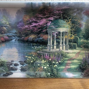 May include: Rectangular serving tray featuring a serene landscape painting. The artwork depicts a river flowing through a lush garden with a gazebo, surrounded by trees and flowers in shades of green, purple, and pink. The tray has a white border.