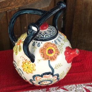 May include: A white ceramic teapot with a floral design featuring orange, yellow, and blue flowers. The teapot has a red spout and a dark blue handle and lid. The lid is topped with a small red knob.