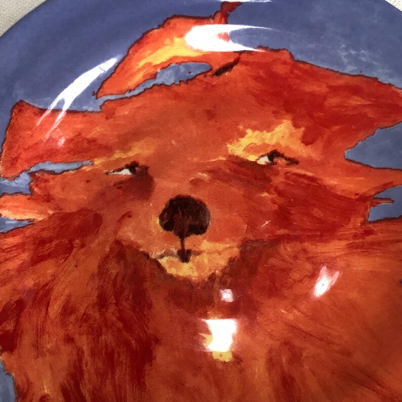May include: A ceramic plate featuring a hand-painted fox face design. The fox is rendered in shades of orange and red, with a dark brown nose and small black eyes. The background is a solid blue.