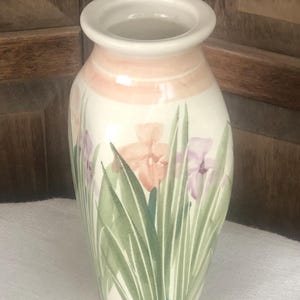 May include: A ceramic vase with a cream-colored base and a pink band near the top. The vase is decorated with green leaves and watercolor-style iris flowers in shades of pink and purple. The vase is approximately 8 inches tall.