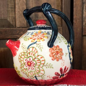 May include: A ceramic teapot with a white base, adorned with colorful floral patterns in red, orange, green, and yellow. It features a red spout and lid, and a dark blue handle. The teapot sits on a red surface.