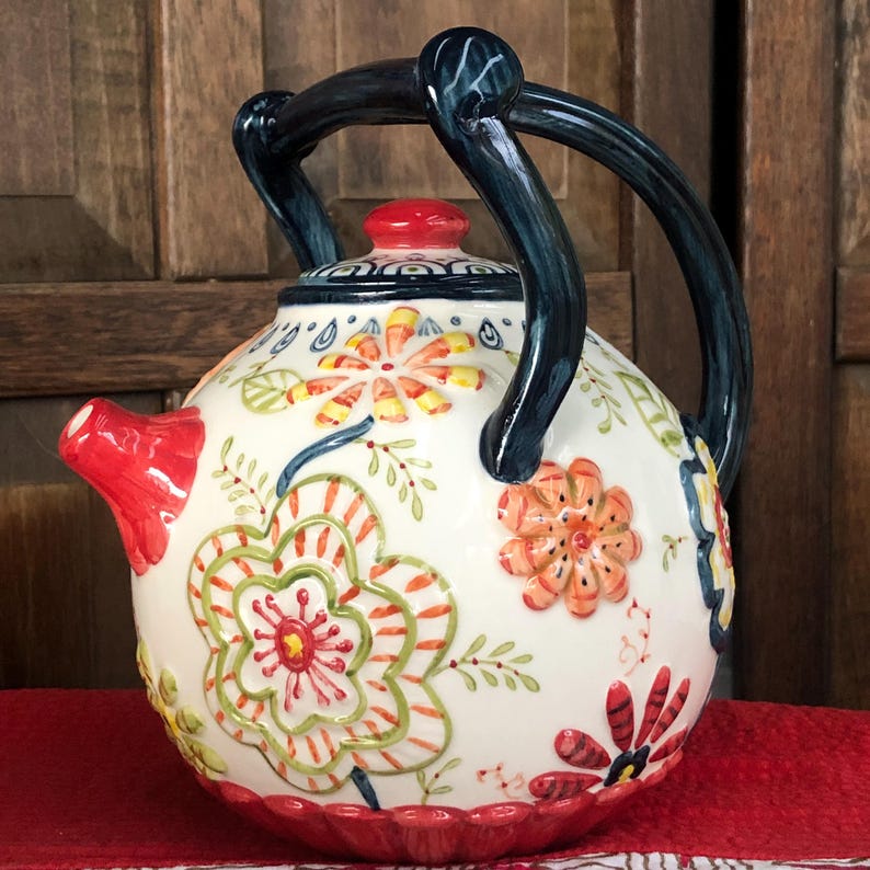 May include: A ceramic teapot with a white base, adorned with colorful floral patterns in red, orange, green, and yellow. It features a red spout and lid, and a dark blue handle. The teapot sits on a red surface.