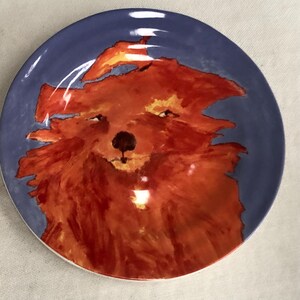 May include: A round ceramic plate featuring a hand-painted illustration of a red-orange dog-like creature. The plate has a blue rim and a white edge. The dog's face is centered, with a black nose and expressive eyes.
