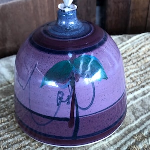 May include: A handcrafted ceramic oil lamp with a purple and maroon color scheme. The lamp features a stylized leaf design in teal and black. The lamp has a white wick and is approximately 4 inches tall.