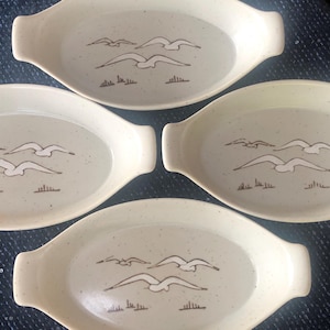 May include: Four oval-shaped, off-white ceramic dishes with handles. Each dish features a brown illustration of three seagulls in flight above a line of grass. The dishes have a speckled texture.