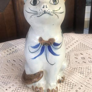 May include: A ceramic cat figurine with a white base, brown accents, and blue details. The cat has black eyes, whiskers, and a bow. The figurine is approximately 6 inches tall.