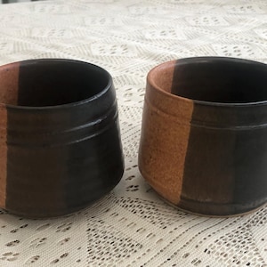 May include: Two ceramic mugs with a brown and tan glaze. The mugs have a vertical stripe design, with one side brown and the other tan.