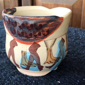 May include: A cream-colored ceramic mug with a unique hand-painted design. The top features a dark blue and orange band with brown chicken illustrations. The lower portion has brown and blue abstract figures on a cream background. The mug has a rounded base.