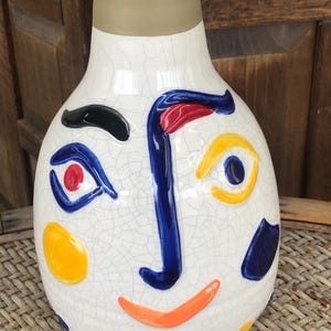 May include: A white ceramic vase with a face design. The vase features a beige neck and is decorated with a blue nose, red and blue eyes, a yellow eye, and an orange smile. The vase has a cracked glaze finish and is adorned with colorful dots.
