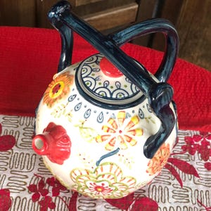 May include: A ceramic teapot with a round body and a dark blue handle. The teapot is decorated with colorful floral patterns in orange, yellow, and green. The spout is red, and the lid has a red knob.