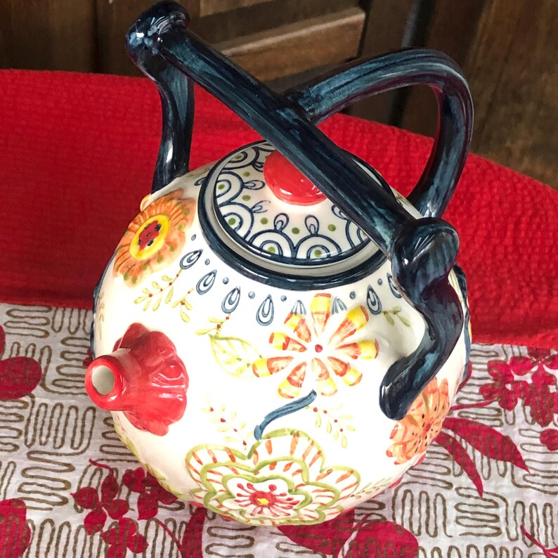 May include: A ceramic teapot with a round body and a dark blue handle. The teapot is decorated with colorful floral patterns in orange, yellow, and green. The spout is red, and the lid has a red knob.