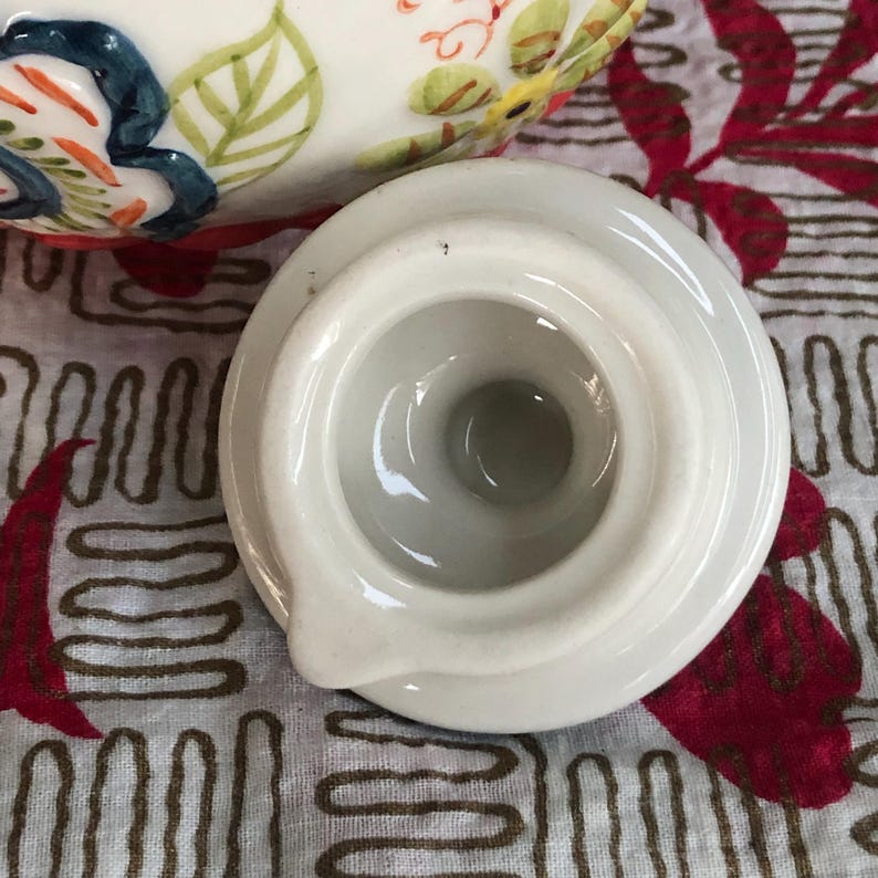 May include: A white ceramic lid with a circular design and a small spout. The lid is positioned next to a portion of a decorated ceramic piece with colorful floral and leaf patterns. The background features a patterned fabric.