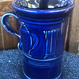 May include: A cobalt blue ceramic mug with a handle. The mug features embossed designs, including a teapot and vertical lines. The mug has a glossy finish and a wide rim. The mug is approximately 5 inches tall.