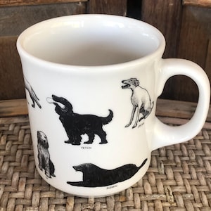 May include: A white ceramic mug featuring black ink illustrations of various dog breeds in different poses. The designs include dogs labeled with the commands Fetch, Speak, and Down, showcasing detailed line work and distinct canine silhouettes.
