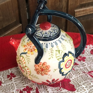May include: A decorative ceramic teapot with a white base, adorned with colorful floral patterns in orange, yellow, and green. It features a red lid and spout, and a dark blue handle. The teapot is placed on a red and white patterned surface.