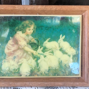 May include: A framed print featuring a young girl in a pink dress feeding a group of white rabbits. The artwork has a vintage aesthetic, with a green and blue background. The frame is made of light brown wood.