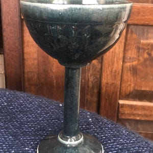 May include: A teal-colored ceramic goblet with a wide bowl and a slender stem. The bowl has a textured pattern around the rim. The base is round with a white edge. The goblet is displayed against a wooden background.