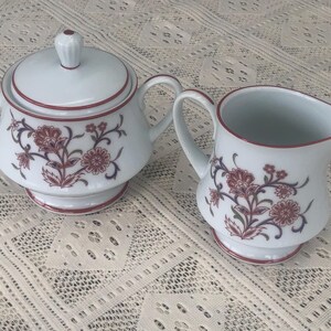 May include: A white sugar bowl and creamer set with a floral design in shades of red and brown. The set has a red rim and a white background.