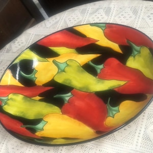 May include: Oval ceramic serving platter with a black background and a colorful chili pepper design. The peppers are red, yellow, and green, with green stems. The platter is on a white lace tablecloth.