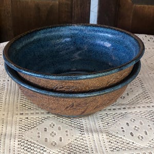 May include: Two handmade ceramic bowls stacked on top of each other. The bowls have a brown exterior with leaf-like patterns and a deep blue interior. The rims are a darker blue. The bowls are sitting on a white lace tablecloth.