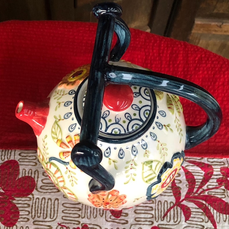 May include: A decorative ceramic teapot with a floral design in orange, yellow, and green on a cream background. The teapot has a red spout and a dark blue handle. The lid features a red knob and a blue and white pattern.