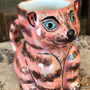 May include: A ceramic mug shaped like a seated cat with a pink base color and dark brown stripes. The cat features large blue eyes, a black nose, and painted whiskers. A curved handle extends from the back, and the interior of the mug is white.